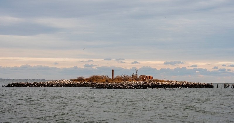 Small island with ruins and vegetation surrounded by water under a cloudy sky.
