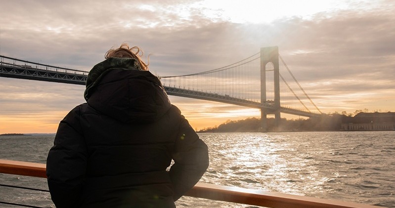 Person in jacket on deck, bridge and sunset over water in background.
