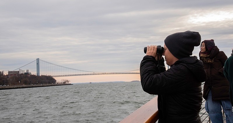 Person on a boat using binoculars to view a distant bridge over water.