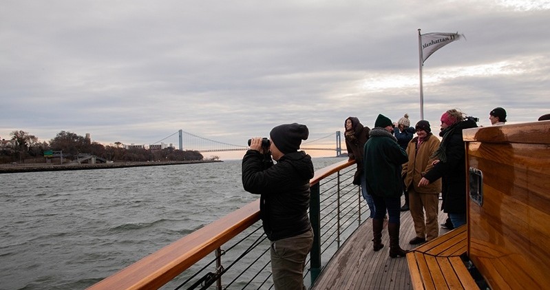 People on a boat deck, one using binoculars, with a suspension bridge and cloudy sky in the background.