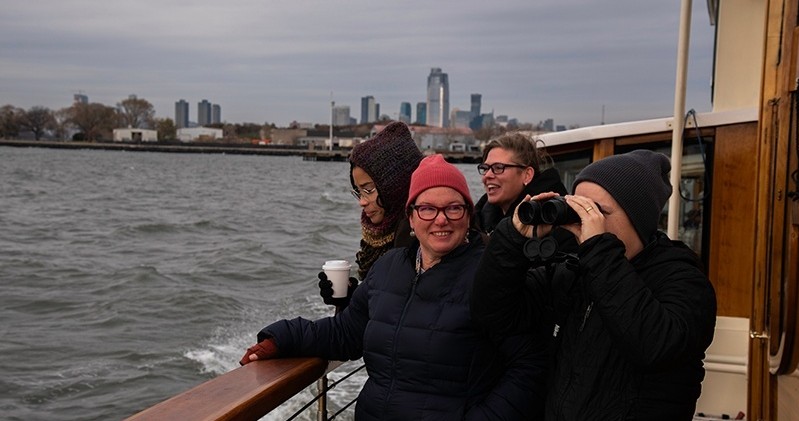 Group of people on a boat, one using binoculars, with city skyline in the background.