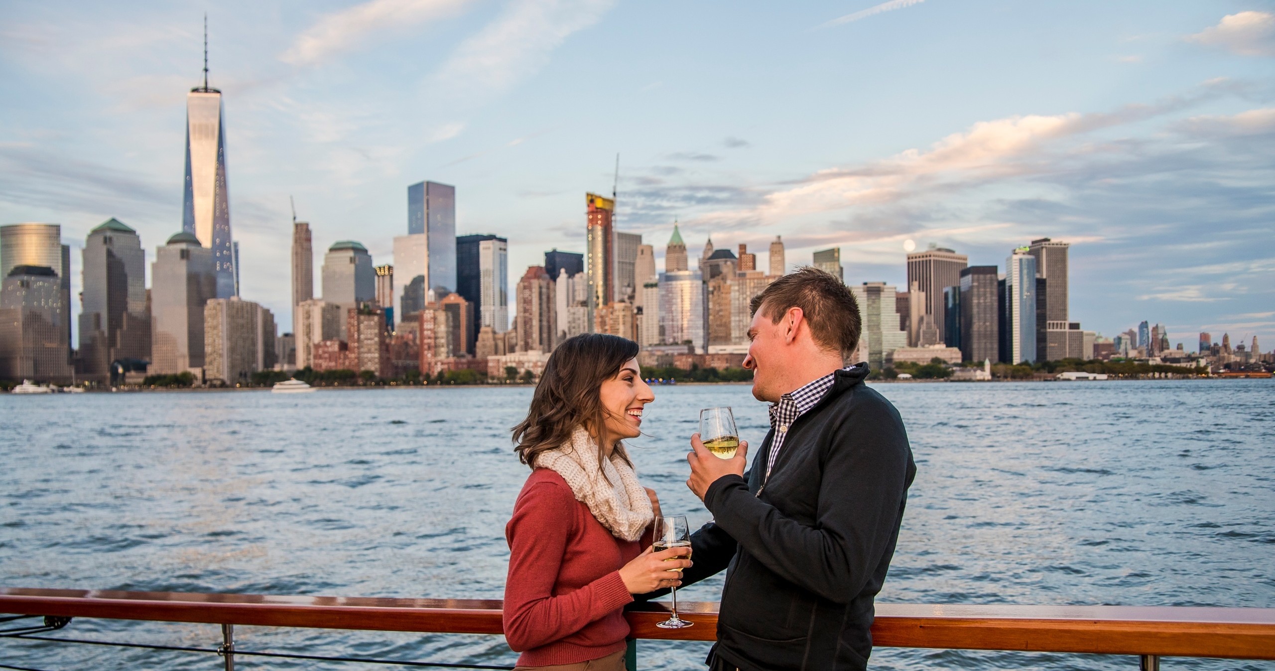 Couple holding drinks on a boat with a city skyline in the background.