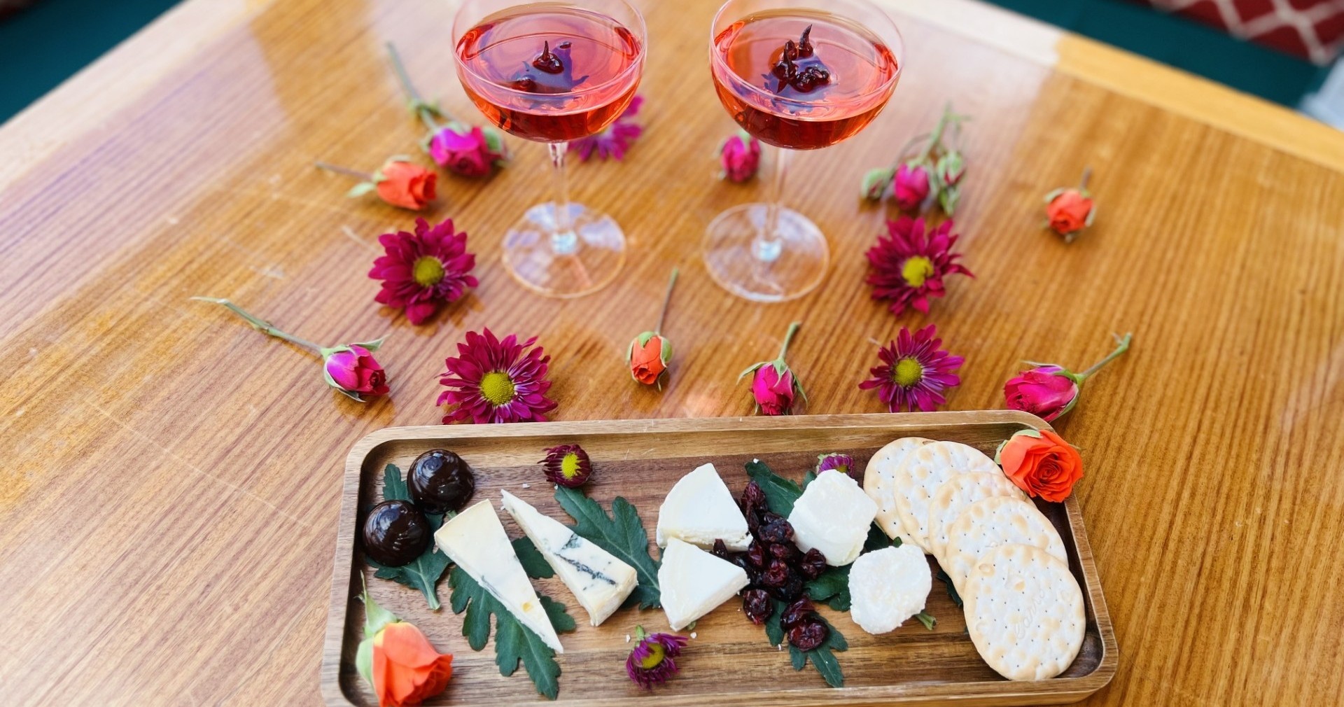 Two glasses of pink drink, cheese, crackers, and flowers on a wooden table.