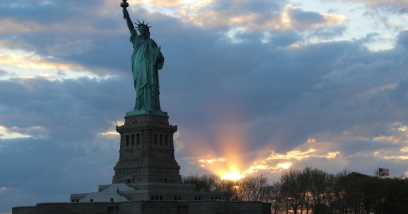 Statue of Liberty silhouetted against a sunset sky with sun rays peeking through clouds.
