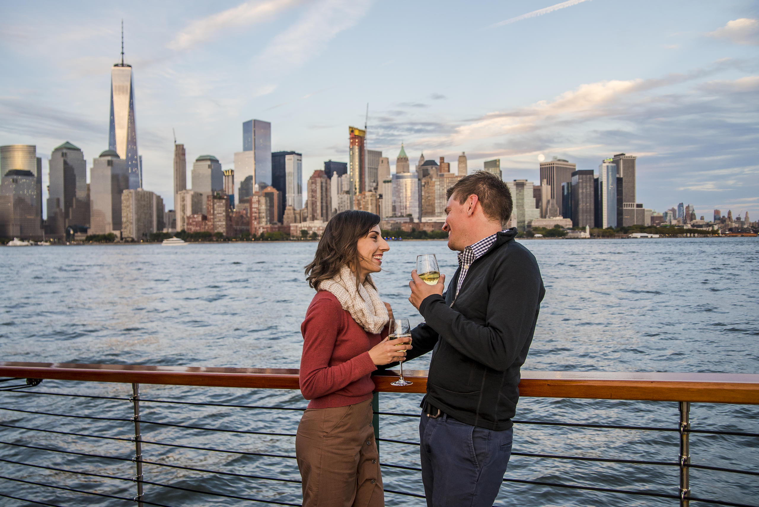 Couple on a boat with drinks, New York City skyline in the background.