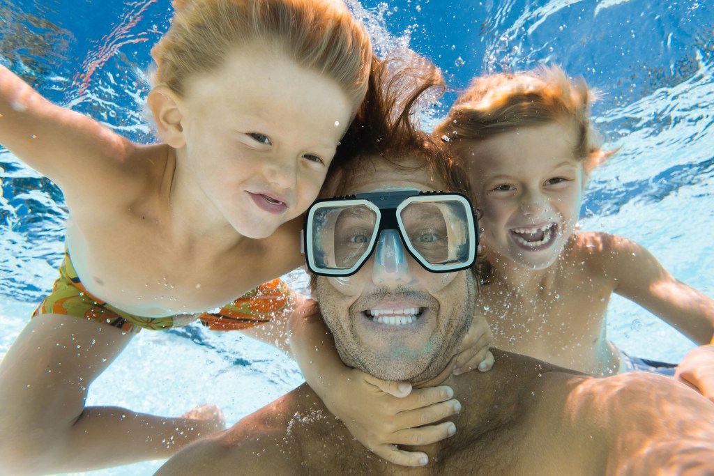 Three people underwater, smiling, one adult with goggles and two children.