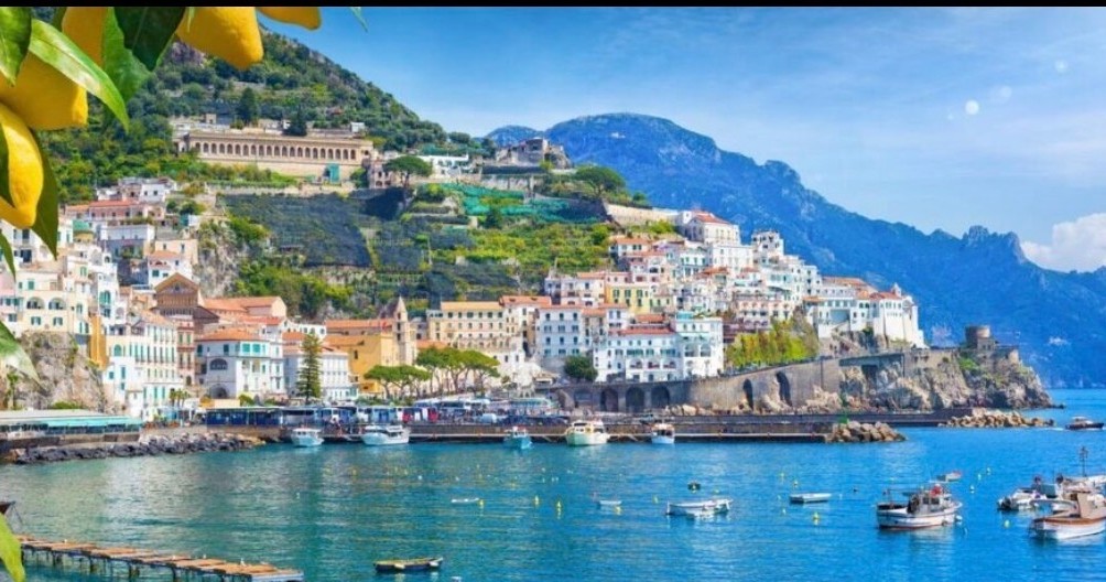 Coastal town with colorful buildings on a hillside, blue sea, boats, and mountains.