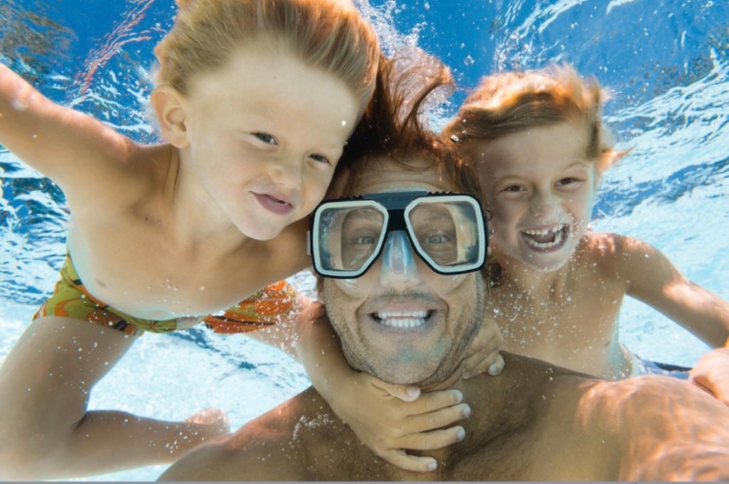 Man with goggles and two children smiling underwater in a pool.