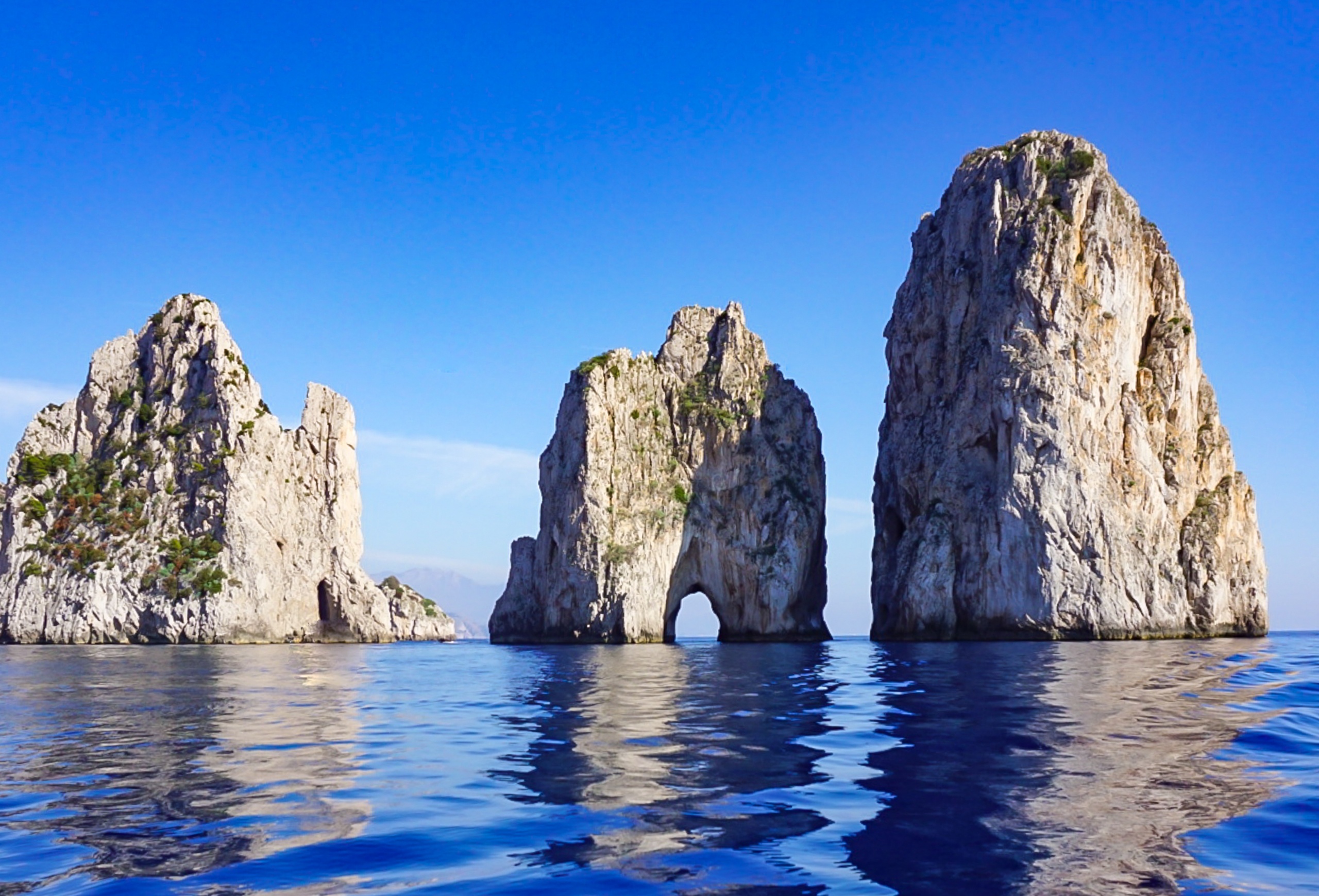 Three rocky sea stacks rise from blue ocean under clear sky.