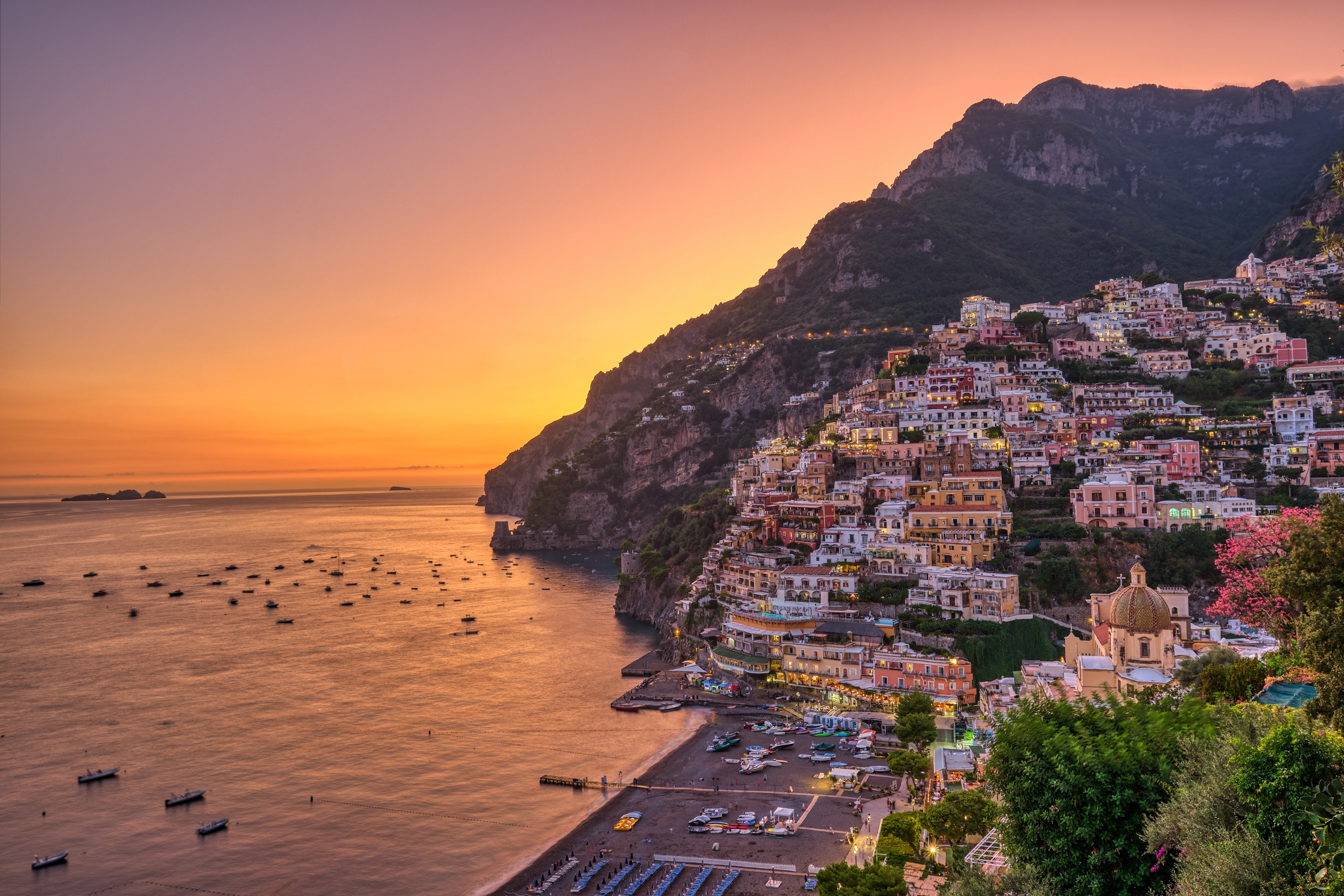 Sunset view of coastal town with colorful buildings on a hillside and boats in the water.