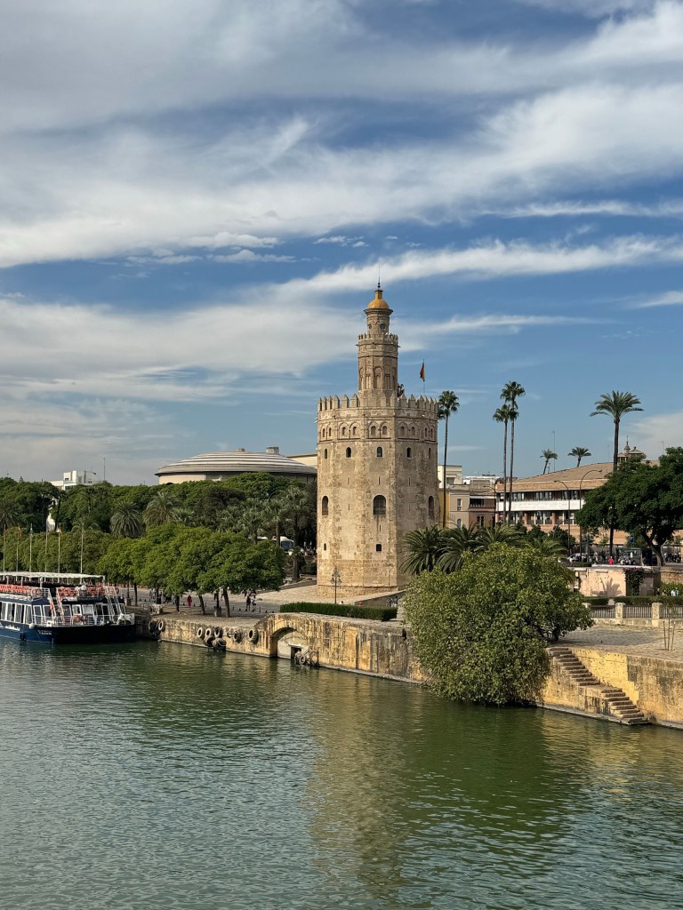 Historic stone tower by a river with boats and palm trees on a sunny day.
