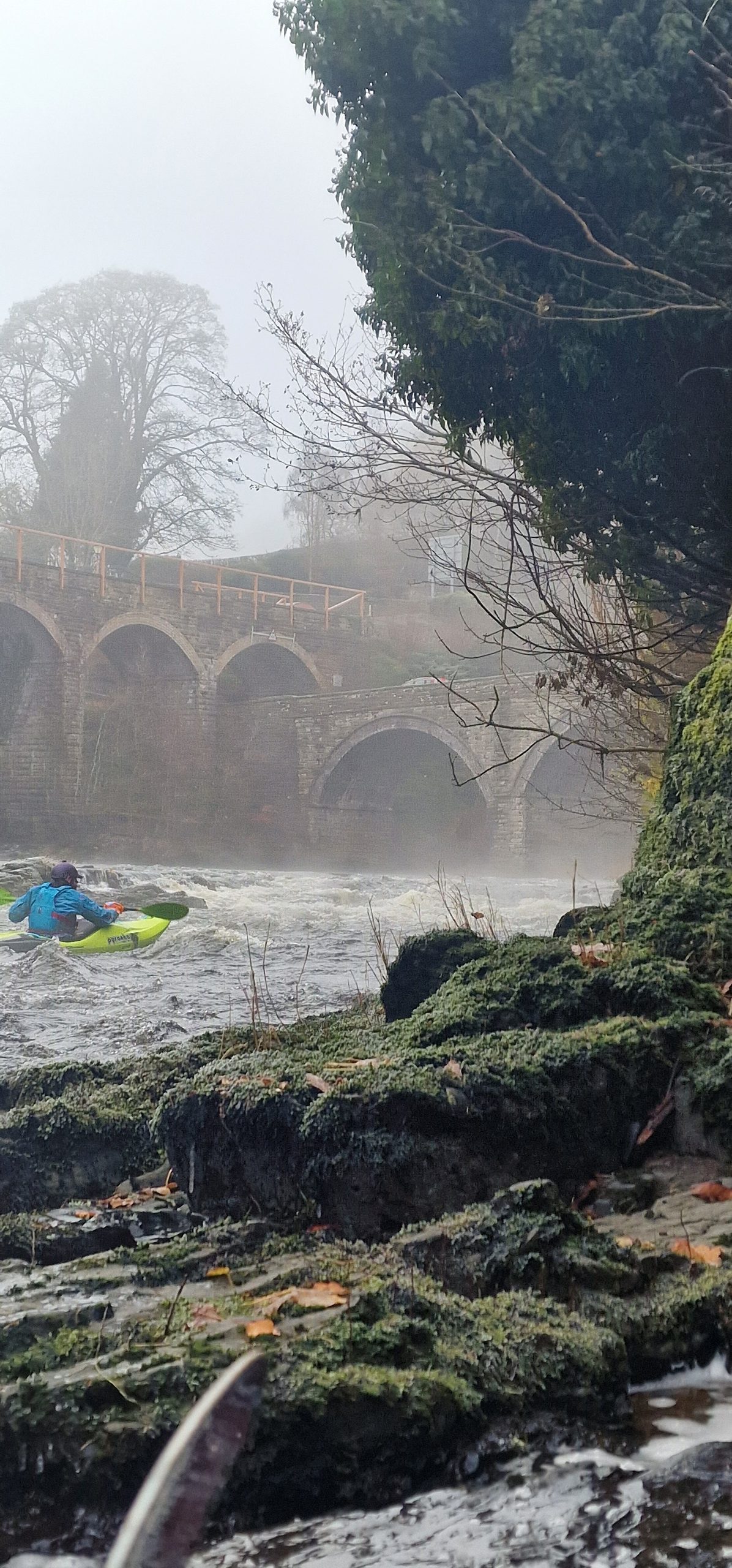 Kayaker on a misty river near an arched stone bridge.