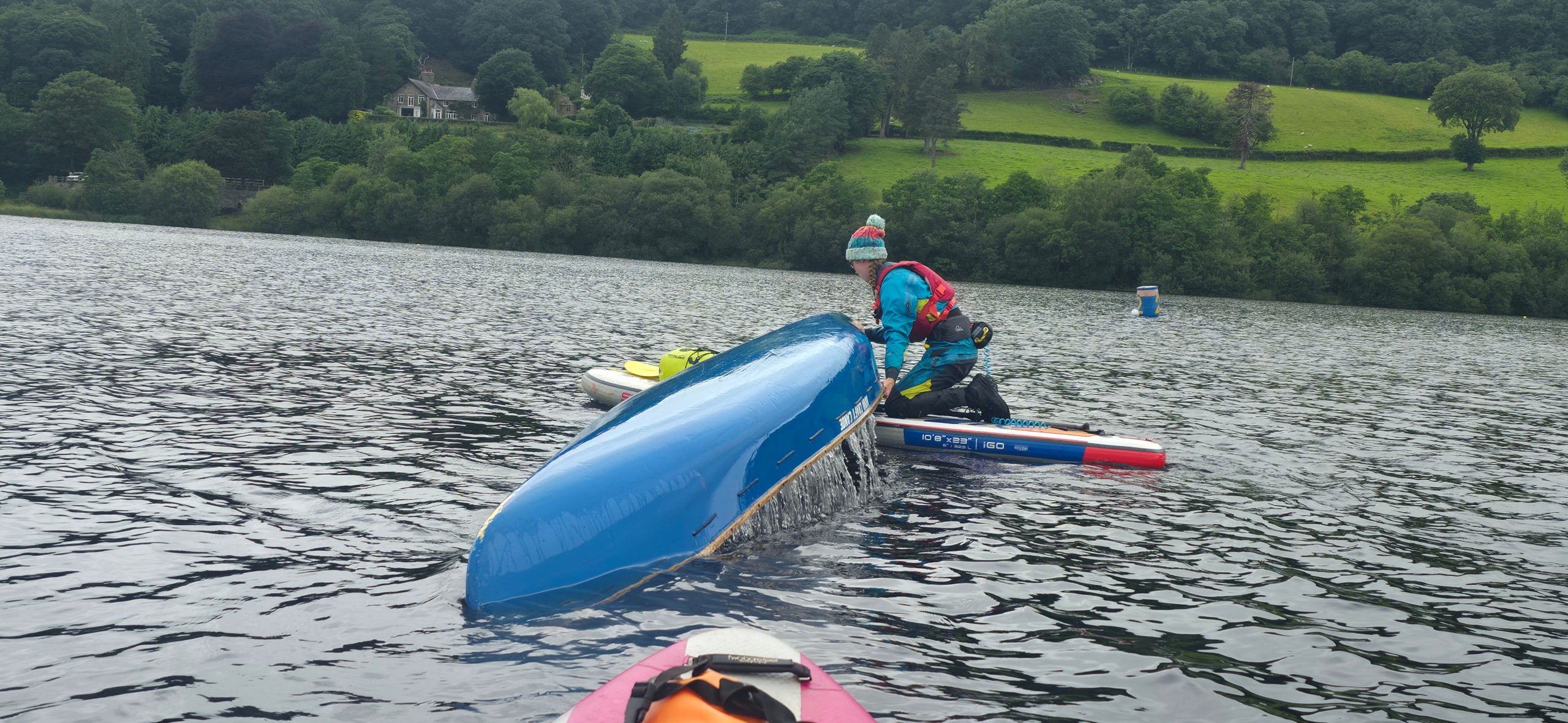 Person in a red kayak assisting an overturned blue kayak on a lake.