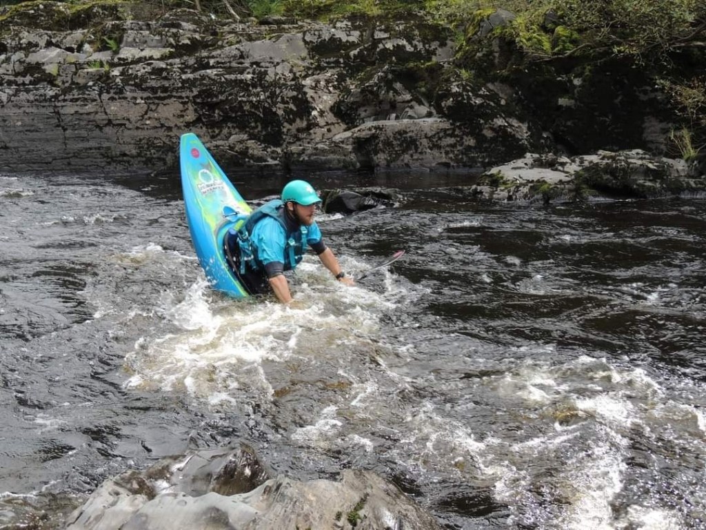 Person kayaking through rocky river rapids wearing a teal helmet and life jacket.