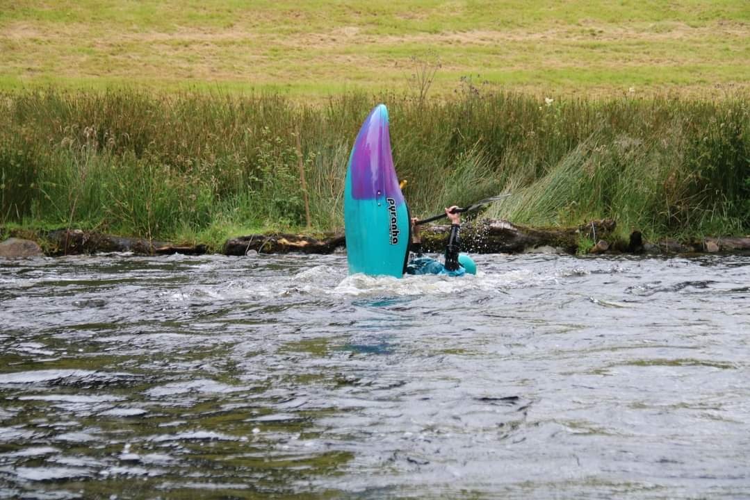 Kayaker vertically balancing in river near grassy bank.