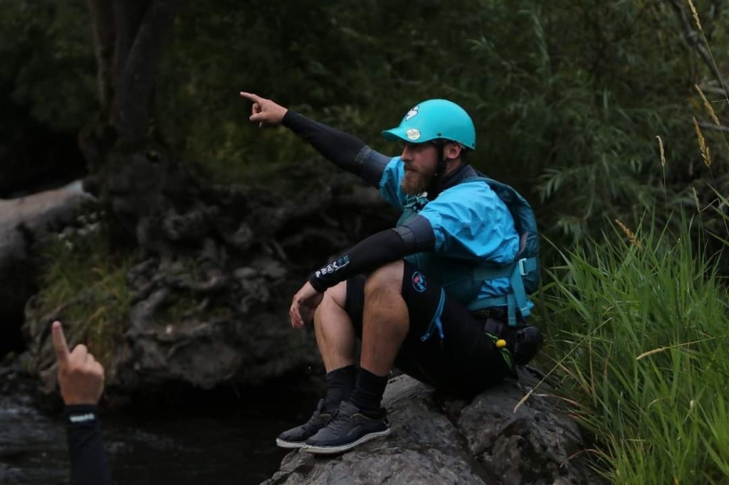 Person in blue gear pointing while sitting on rock by river.