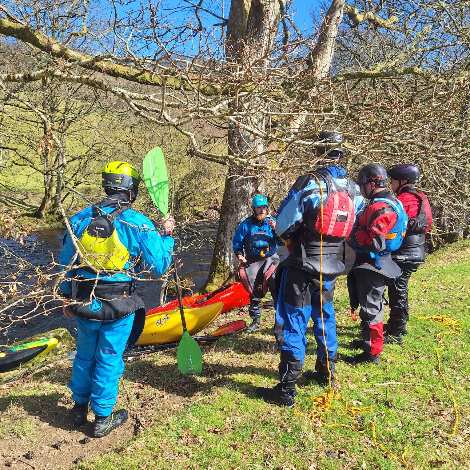 Group of kayakers in colorful gear gather by a riverbank with kayaks.