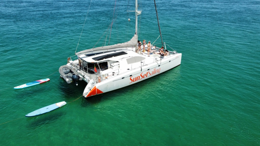 Catamaran named 'Sunset' with people sunbathing, paddleboards floating nearby in turquoise water.