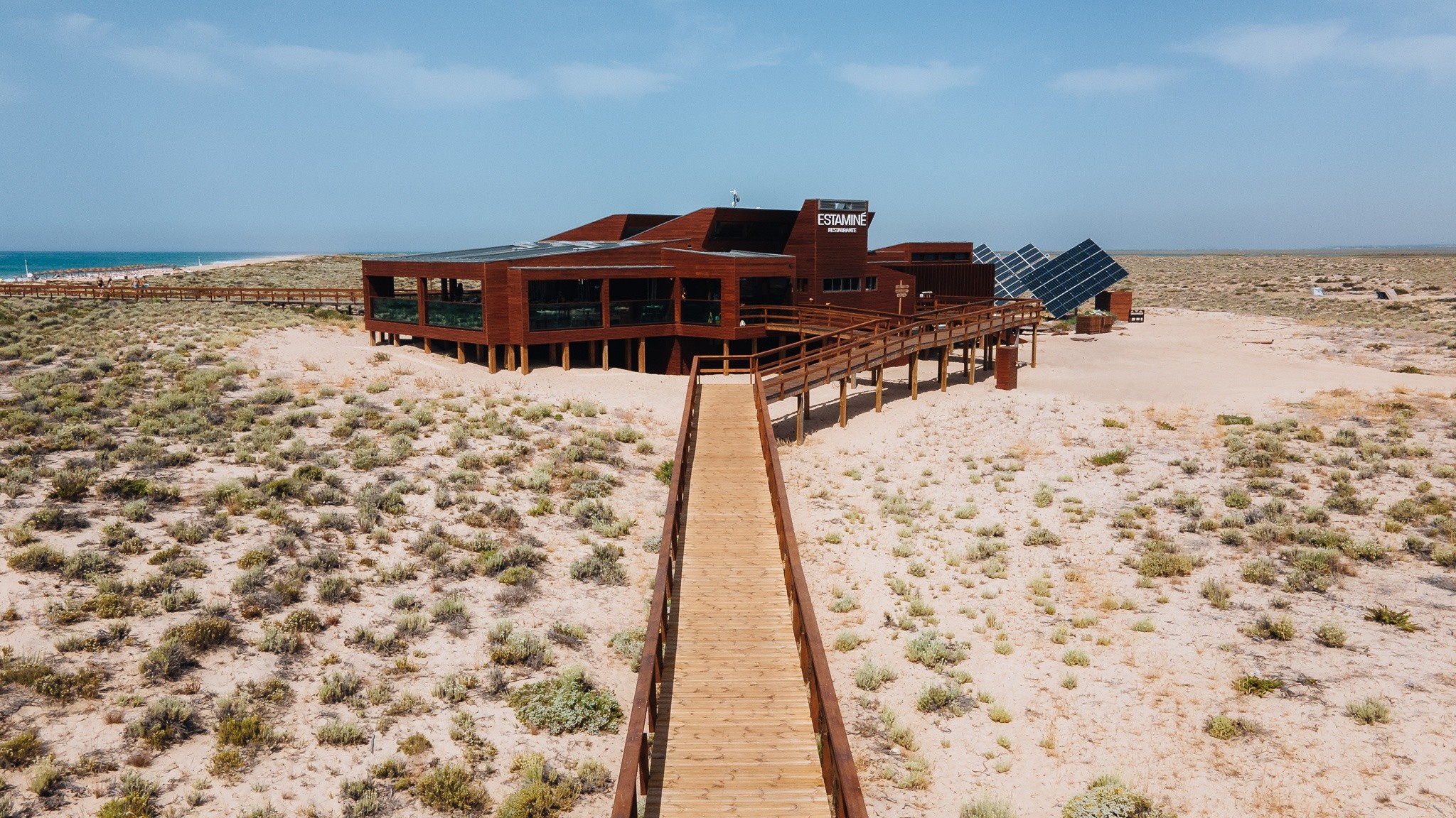 Wooden building with solar panels in desert, long walkway leading to it.