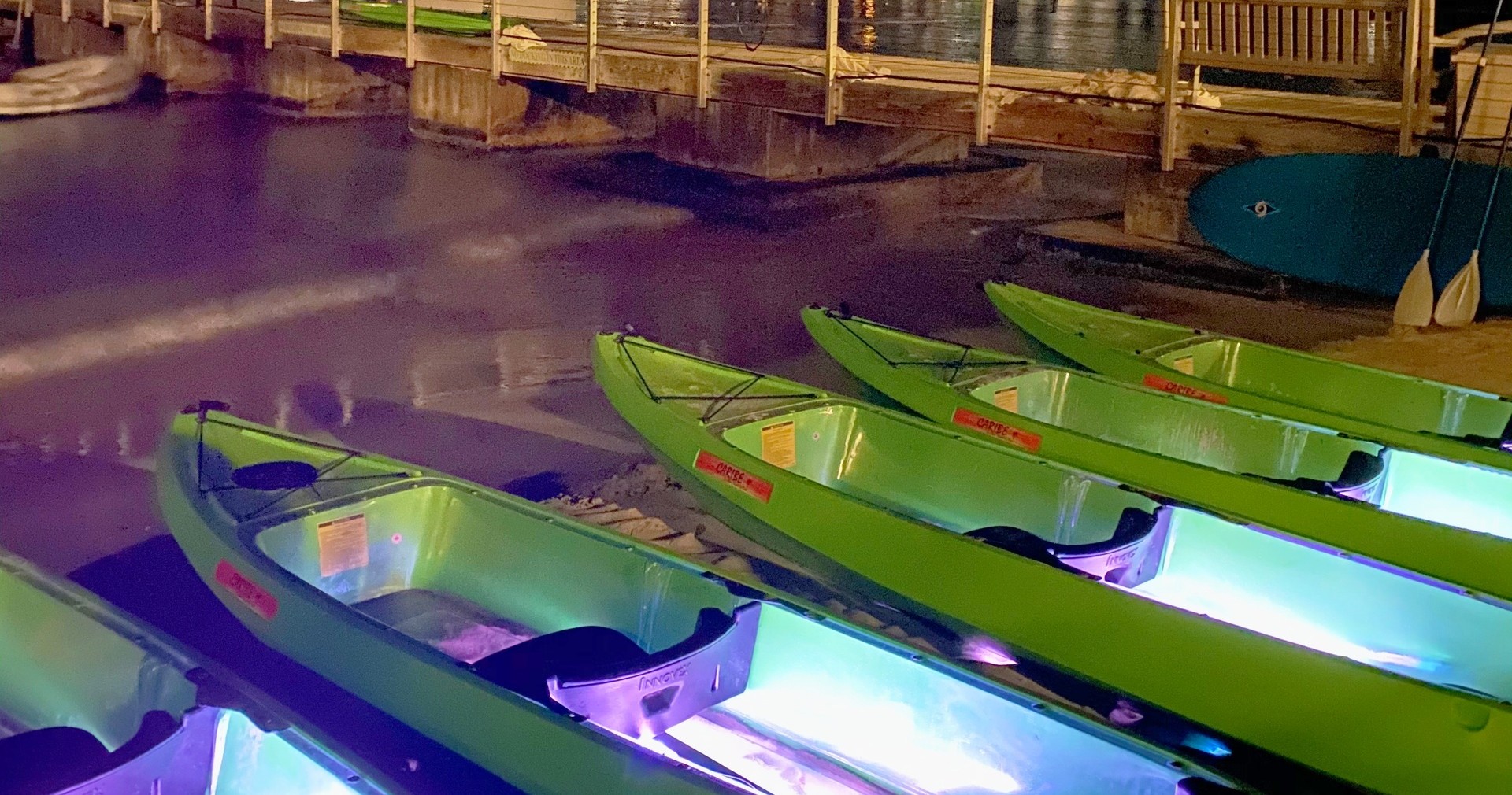 Row of illuminated green kayaks on a beach at night with a wooden pier in the background.