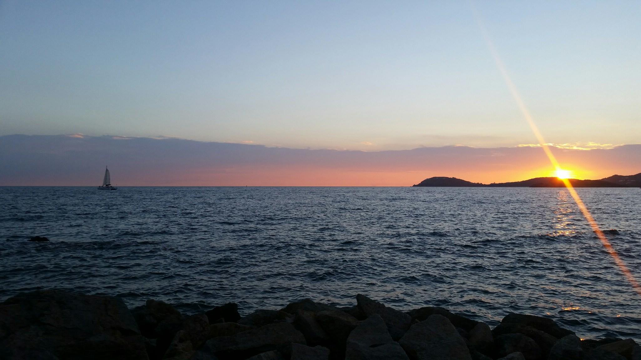 Sunset over ocean with sailboat and rocky shore.