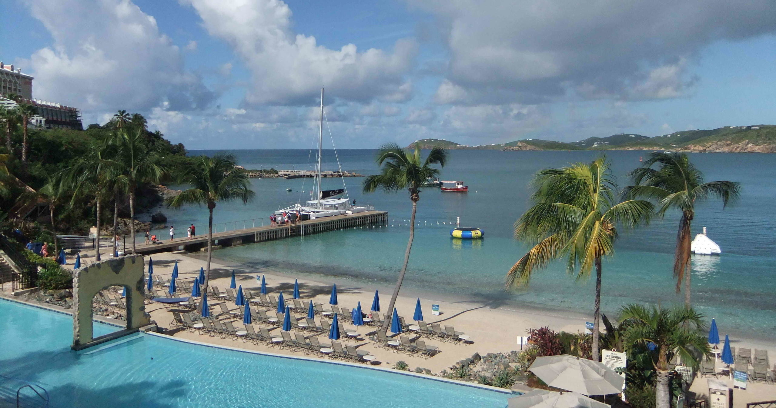 Tropical beach with palm trees, umbrellas, pool, and boats on a sunny day.