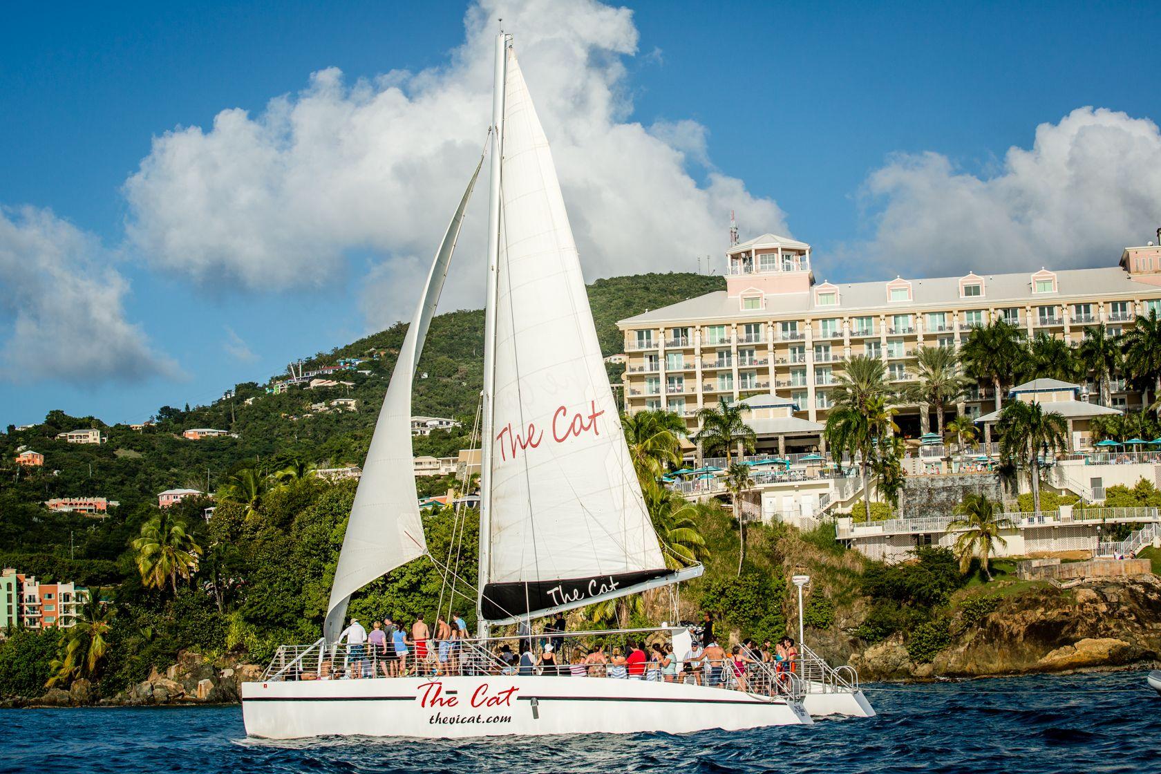 Catamaran sailing near a tropical resort with people onboard.