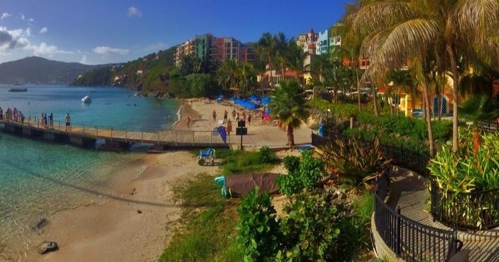 Sunny beach with colorful buildings, palm trees, and people near turquoise water.