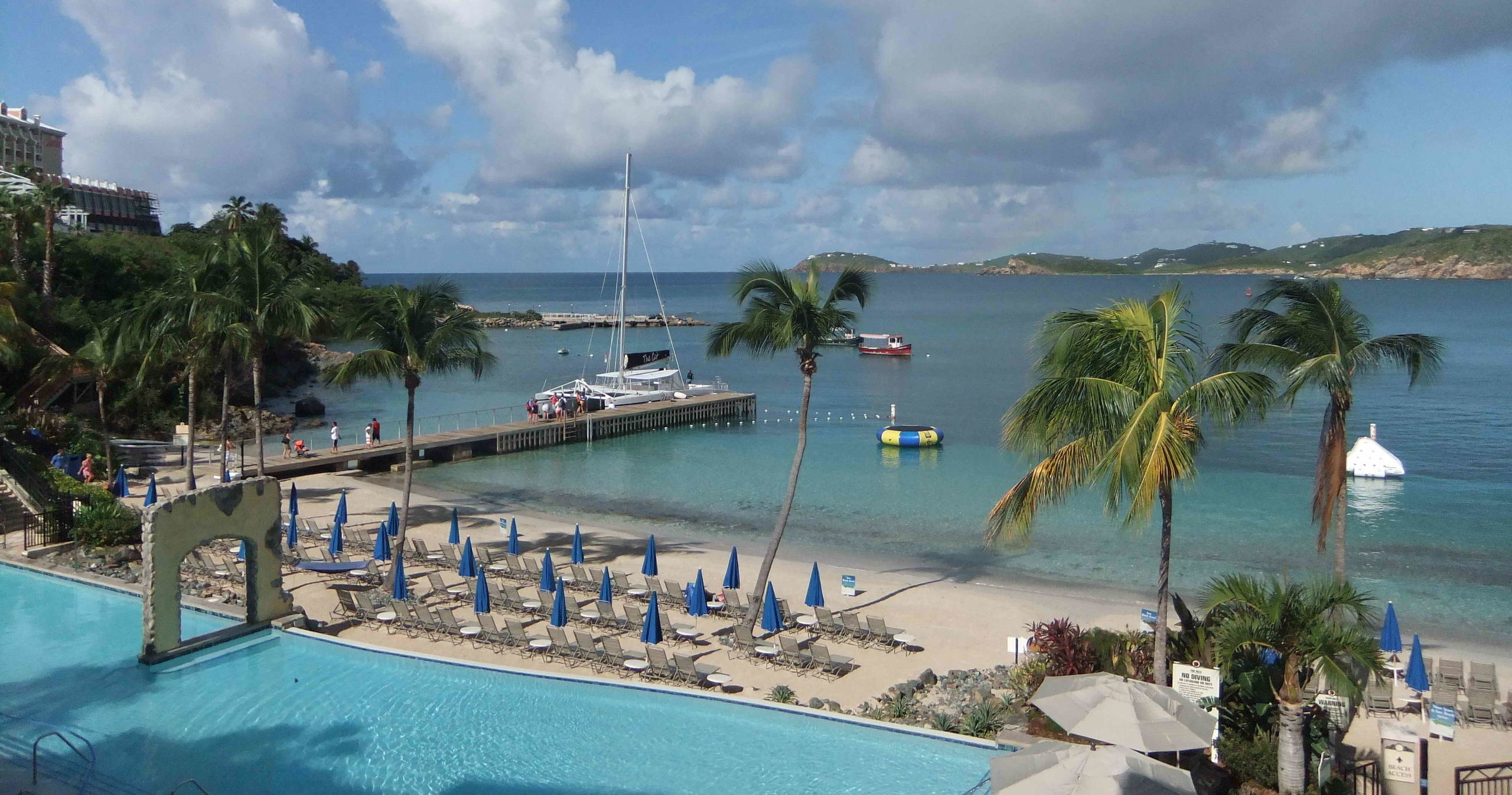 Tropical beach scene with palm trees, blue umbrellas, and boats on the water.