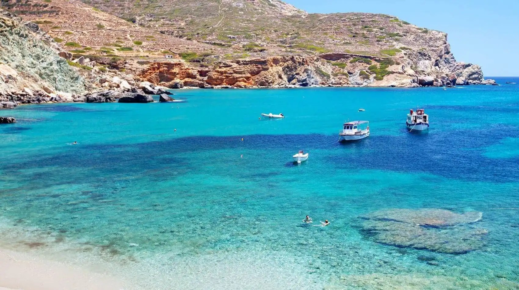 Boats on clear turquoise water near rocky coast under blue sky.