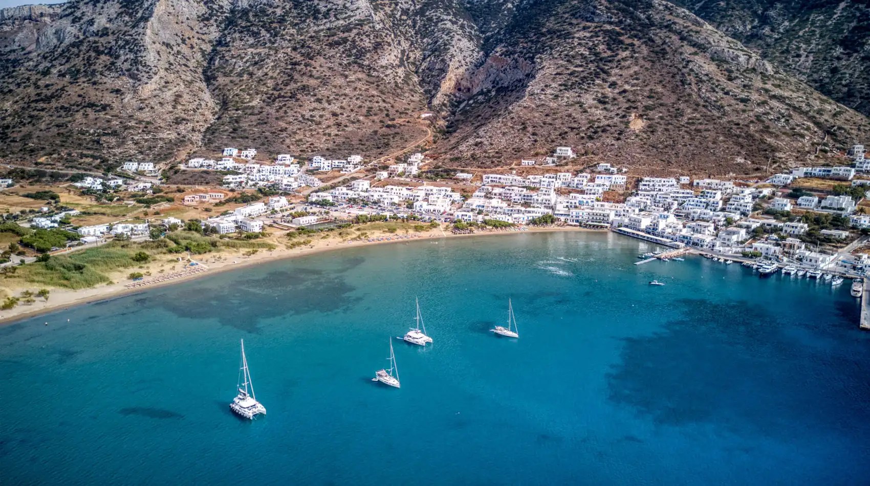 Coastal town with sailboats in blue bay, surrounded by hills.