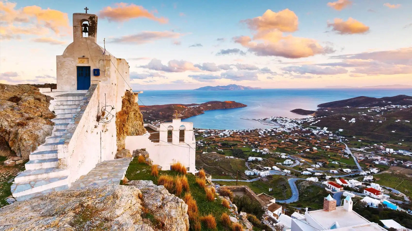 Greek hillside church overlooking coastal town and sea at sunset.