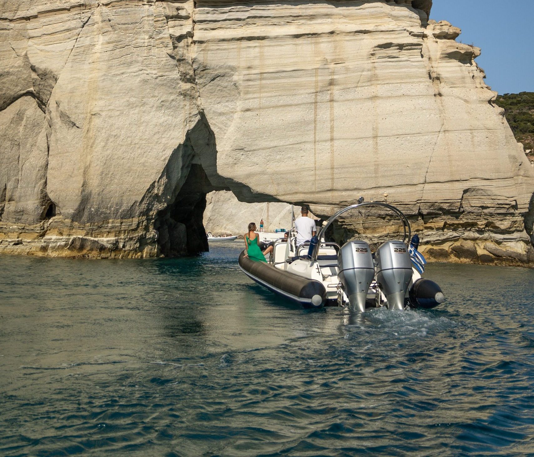 Small boat approaching rock formation arch over blue water.