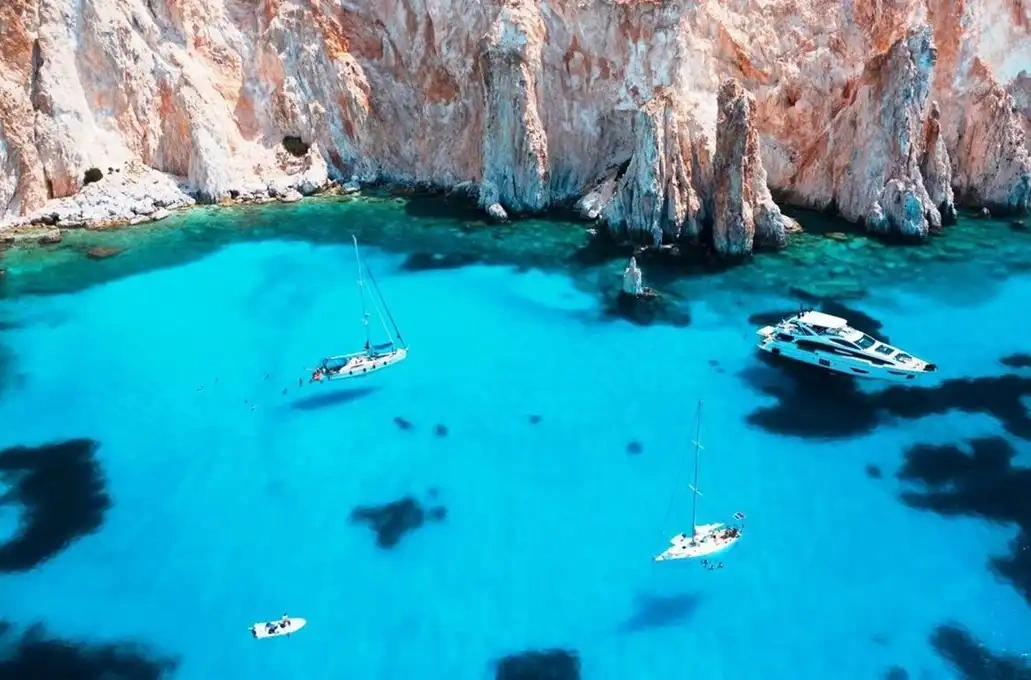 Sailboats and a yacht in clear blue water near rugged cliffs.