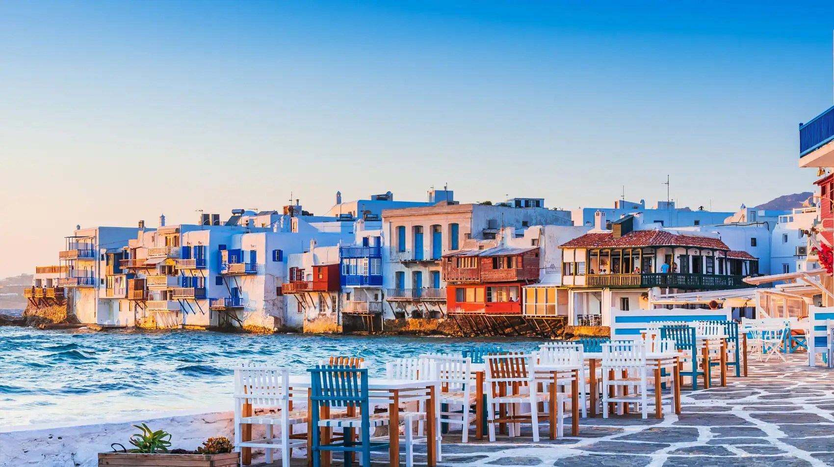 Colorful seaside buildings with white chairs and tables on a stone walkway during sunset.