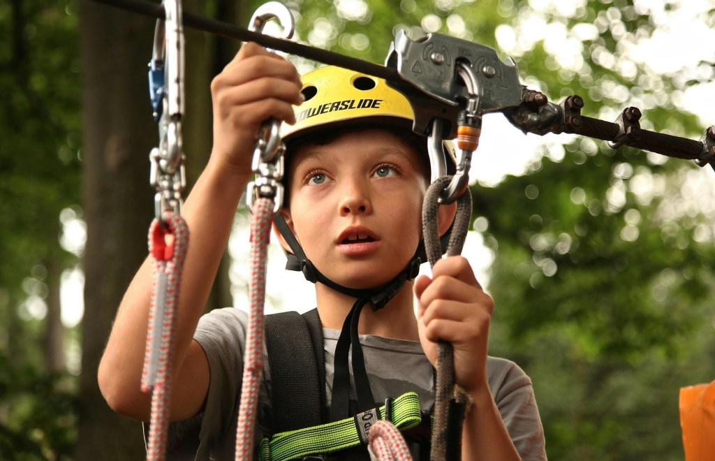 Child wearing safety gear, adjusting a harness on a ropes course in a forest.