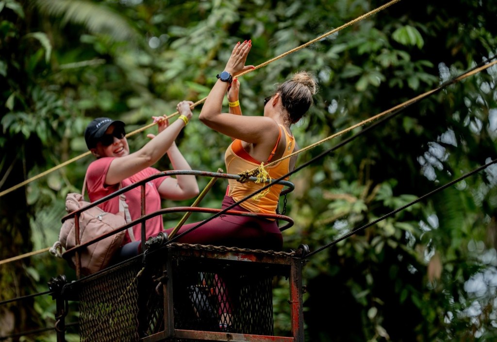 Two women high-five on a zip line platform in a forest.