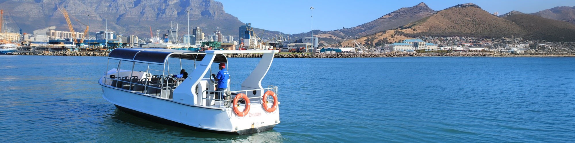 Small boat on a calm waterway with mountains and a cityscape in the background.