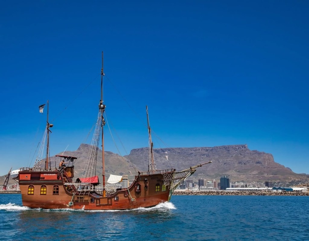 Old wooden ship sailing near mountains under clear blue sky.